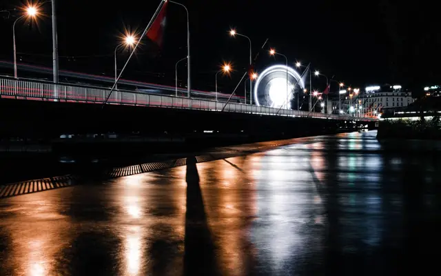 Stunning night view of Geneva bridge with illuminated ferris wheel, beautifully reflected in the wet pavement.