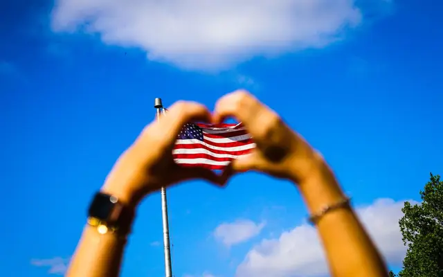 Hands form a heart shape around an American flag against a vibrant blue sky, symbolizing patriotism and freedom.