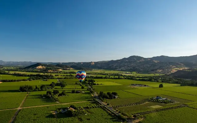 Colorful hot air balloon flying above Napa Valley vineyards under a clear blue sky.