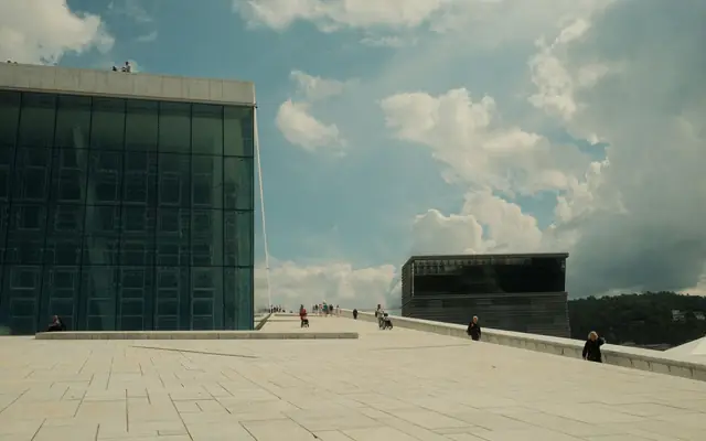 Wide angle view of Oslo Opera House with people on its sloped roof under a cloudy sky.